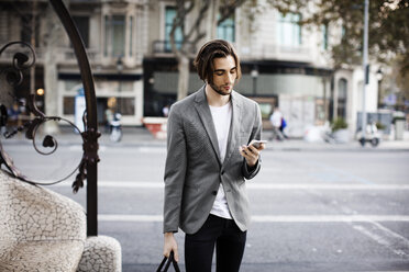 Man with bag using mobile phone while standing on city street - CAVF33903