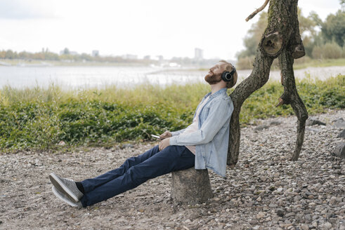 Germany, Duesseldorf, man with smartphone and headphones listening music in nature - KNSF03660