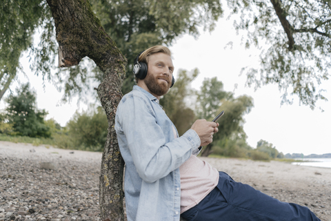 Deutschland, Düsseldorf, Mann hört Musik mit Smartphone und Kopfhörer am Strand, lizenzfreies Stockfoto