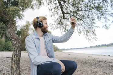 Deutschland, Düsseldorf, Mann mit Kopfhörer macht Selfie am Strand mit Smartphone - KNSF03663