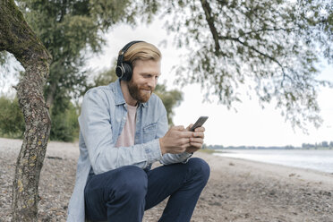 Deutschland, Düsseldorf, lächelnder Mann mit Kopfbedeckung und Smartphone am Strand - KNSF03664