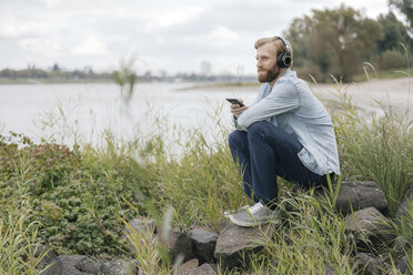 Germany, Duesseldorf, man listening music with headphones and smartphone in nature - KNSF03665