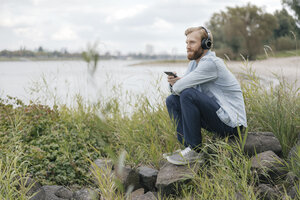 Deutschland, Düsseldorf, Mann hört Musik mit Kopfhörer und Smartphone in der Natur - KNSF03665