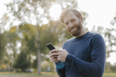 Porträt eines lächelnden Mannes mit Mobiltelefon in einem Park - KNSF03680