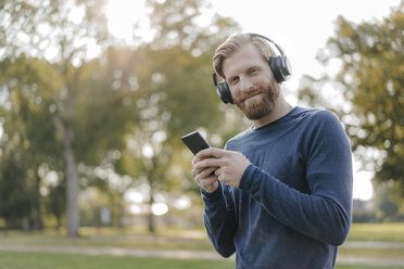 Portrait of smiling man with cell phone listening music with headphones in a park - KNSF03681