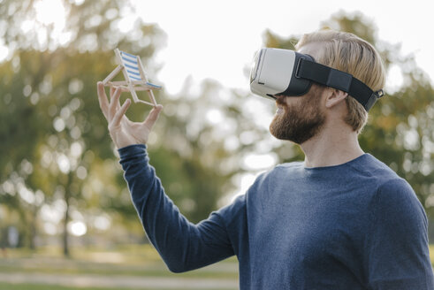 Mann in herbstlichem Park mit Virtual-Reality-Brille betrachtet Miniatur-Strandliege - KNSF03684