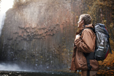 Male hiker carrying backpack while looking at waterfall - CAVF33969