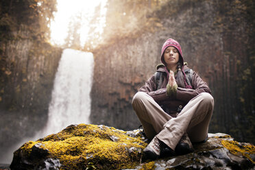 Frau meditiert auf einem Felsen mit Wasserfall im Hintergrund - CAVF33972