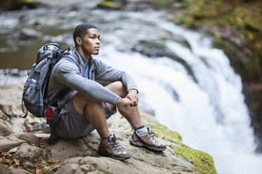 Nachdenklicher männlicher Wanderer sitzt auf einem Felsen am Wasserfall - CAVF33981