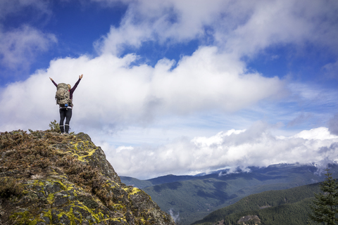 Niedriger Blickwinkel auf eine Frau, die mit erhobenen Armen auf einem Berg vor einem bewölkten Himmel steht, lizenzfreies Stockfoto