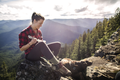 Frau mit Hund auf einem Felsen sitzend und Ukulele spielend an einer Bergklippe, lizenzfreies Stockfoto