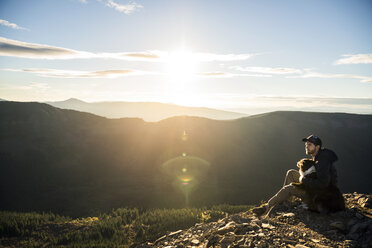 Mann mit Hund auf einer Klippe vor einer Bergkette sitzend - CAVF34023