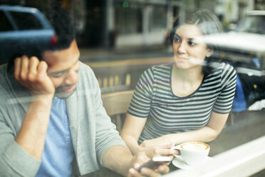 Woman looking at man using smart phone while sitting in cafe seen through glass window - CAVF34037