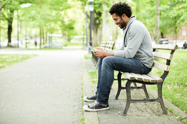 Seitenansicht eines glücklichen Mannes, der sein Smartphone benutzt, während er auf einer Parkbank am Fußweg sitzt - CAVF34047