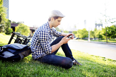 Mann, der sein Smartphone benutzt, während er auf einer Wiese vor einem klaren Himmel sitzt - CAVF34068
