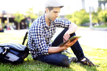 Mann, der ein digitales Tablet benutzt, während er auf einer Wiese an einem sonnigen Tag sitzt - CAVF34071