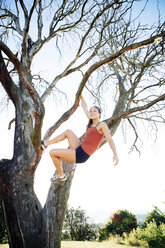 Happy sporty woman hanging from branch against clear sky - CAVF34078