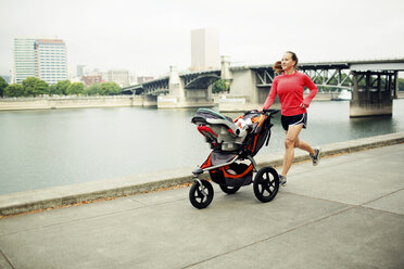 Frau joggt mit Kinderwagen auf Fußweg am Fluss gegen Brücke - CAVF34089