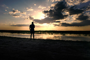 Silhouette man standing at lakeshore against cloudy sky during sunset - CAVF34245