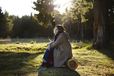 Seitenansicht einer nachdenklichen Frau, die wegschaut, während sie auf einem Baumstamm im Wald sitzt - CAVF34260