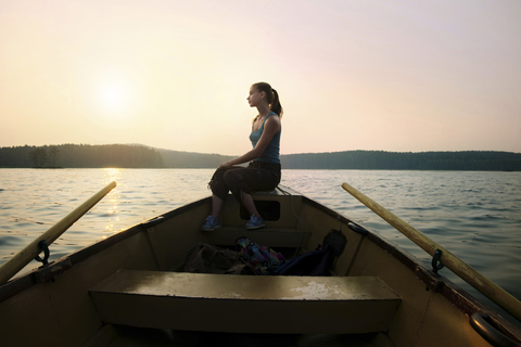 Nachdenkliche Frau auf dem Schiffsbug sitzend im Kanu gegen den klaren Himmel in der Abenddämmerung, lizenzfreies Stockfoto