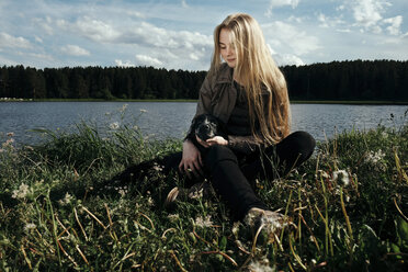 Woman sitting with dog by lake against cloudy sky - CAVF34302