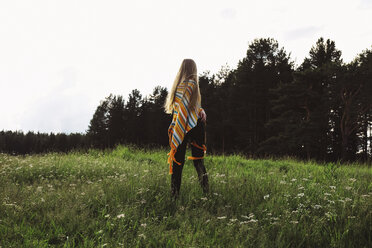 Rückansicht einer Frau im Poncho, die auf einer Wiese vor einem wolkenlosen Himmel steht - CAVF34316