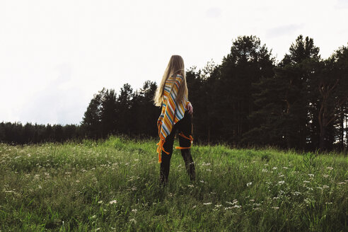 Rear view of woman in poncho standing on grassy field against clear sky - CAVF34316