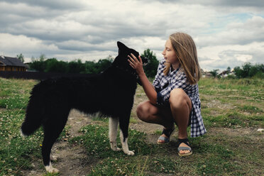 Teenager-Mädchen, das einen Hund streichelt, während es auf einem Feld vor stürmischen Wolken kauert - CAVF34356