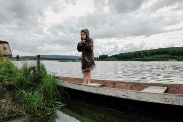 Girl wearing hooded jacket while standing in rowboat on lake - CAVF34359