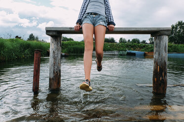 Low section of teenage girl playing with water while sitting on bench over lake - CAVF34362