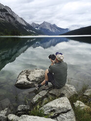 Mann sitzt auf einem Felsen und fotografiert den See mit seiner Kamera - CAVF34389