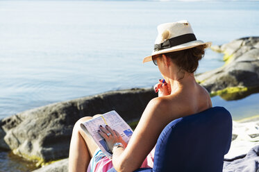 Frau sitzt im Stuhl und liest Zeitung am felsigen Strand - FOLF09222