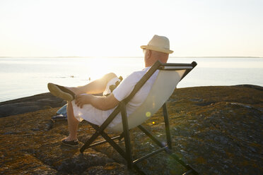 Älterer Mann entspannt sich im Liegestuhl am felsigen Strand - FOLF09226