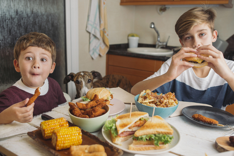 Zwei Jungen mit Hund genießen amerikanisches Essen am Esstisch zu Hause, lizenzfreies Stockfoto