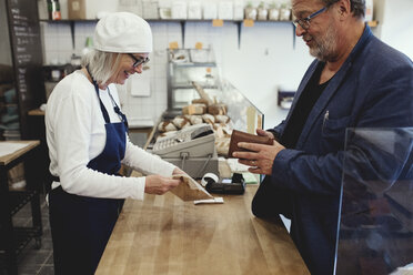 Male customer paying female baker while standing at checkout corner in bakery - MASF00092