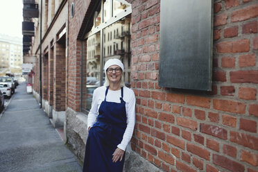 Porträt eines lächelnden Bäckers, der sich an eine Backsteinmauer vor einer Bäckerei lehnt - MASF00099
