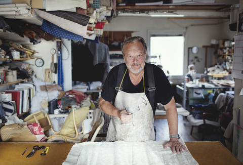 Porträt eines älteren männlichen Besitzers, der mit Stoff an einer Werkbank in einer Werkstatt steht, lizenzfreies Stockfoto