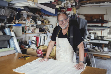 Portrait of smiling male owner standing with fabric at workbench - MASF00112