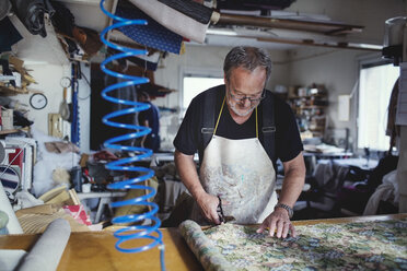 Senior male owner cutting fabric at workbench in workshop - MASF00113