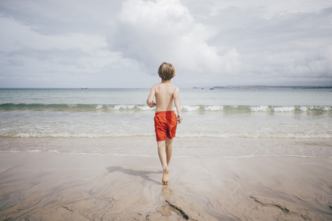 Rückansicht eines Jungen ohne Hemd, der am Strand gegen den Himmel läuft, lizenzfreies Stockfoto