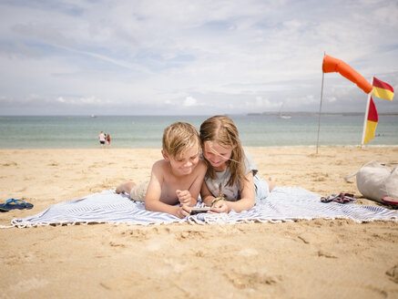 Geschwister teilen sich ein Smartphone, während sie auf einem Handtuch am Strand gegen den Himmel liegen - MASF00136