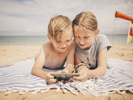 Lächelnde Geschwister, die sich ein Smartphone teilen, während sie auf einem Handtuch am Strand gegen den Himmel liegen - MASF00137