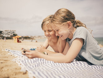 Glückliche Geschwister teilen sich ein Smartphone, während sie am Strand auf einem Handtuch liegen und den Himmel betrachten - MASF00138