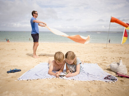 Mann hält Handtuch, während Kinder ihr Smartphone am Strand gegen den Himmel austauschen - MASF00139