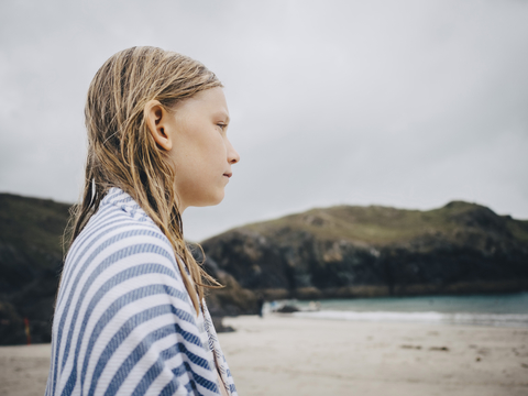 Seitenansicht eines blonden Mädchens, eingewickelt in ein gestreiftes blaues Handtuch, das am Strand vor dem Himmel steht, lizenzfreies Stockfoto