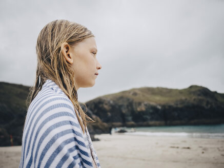 Side view of blond girl wrapped in striped blue towel standing at beach against sky - MASF00143