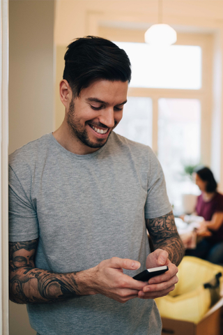 Lächelnder Mann, der ein Mobiltelefon benutzt, während er sich an die Tür seines neuen Hauses lehnt, lizenzfreies Stockfoto