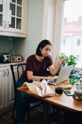 Woman using laptop while holding mobile phone at dining table in kitchen - MASF00171