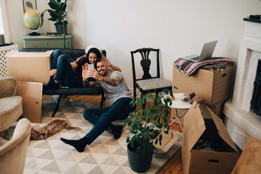 Lächelndes Paar, das während eines Umzugs im Wohnzimmer sitzend über das Mobiltelefon Videogespräche führt - MASF00177
